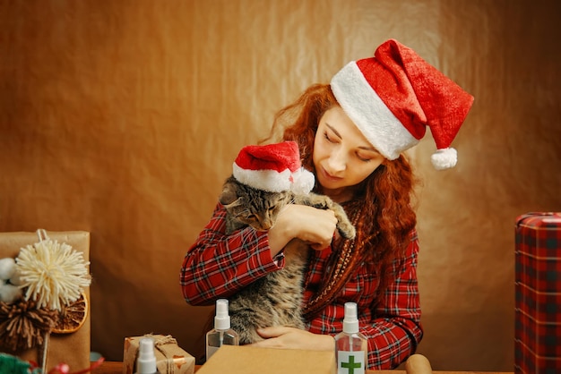 Hermosa chica con sombrero de navidad con gato en sombrero de santa claus junto a la mesa con cajas de regalo y desinfectante ...