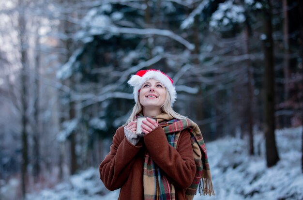 Hermosa chica con sombrero de Navidad con una taza en un bosque nevado