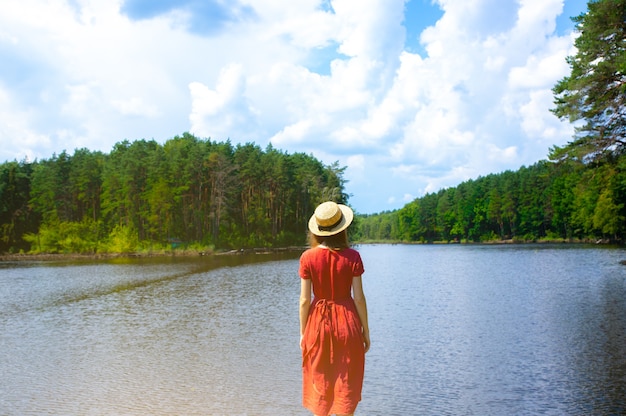 Hermosa chica con sombrero de paja cerca del lago. Increíble naturaleza de verano. Concepto de viaje. Inspiración para la pasión por los viajes. Mujer en vestido rojo de lino. Materiales ecológicos para ropa.