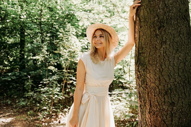 Hermosa chica con sombrero de paja y ropa elegante se encuentra en un parque cerca de un árbol. Una mujer con un vestido blanco cerca de un árbol. Cerrar retrato de niña feliz