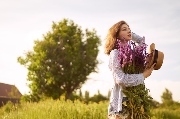 Una hermosa chica con un sombrero con un ramo de lavanda. Ella está vestida con una camisa azul, una camiseta blanca y un sombrero.