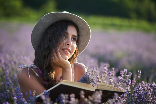 Hermosa chica con sombrero sentada en un campo de lavanda púrpura y leyendo un libro