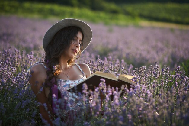 Hermosa chica con sombrero sentado en el campo de lavanda púrpura y leer el libro.