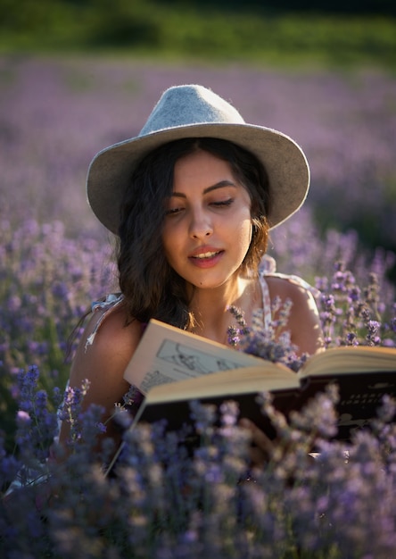 Hermosa chica con sombrero sentado en el campo de lavanda púrpura y leer el libro.