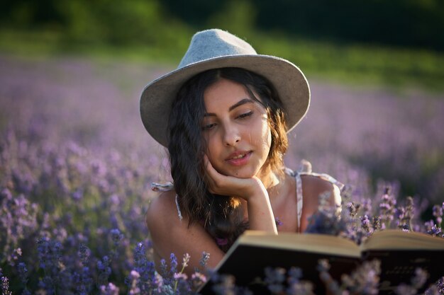 Hermosa chica con sombrero sentado en el campo de lavanda púrpura y leer el libro.