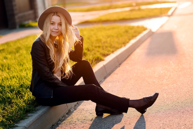 Hermosa chica con un sombrero sentado en el lado de la acera de la carretera al atardecer