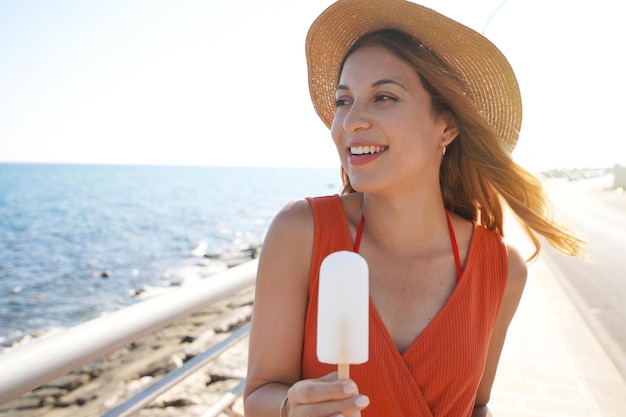 Hermosa chica con sombrero sosteniendo una paleta blanca en la playa mirando hacia un lado