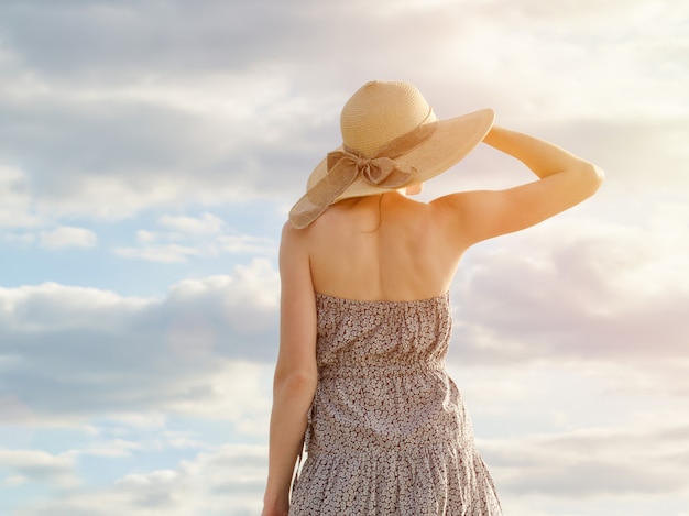 Hermosa chica con sombrero y vestido mirando lejos contra un cielo azul nublado