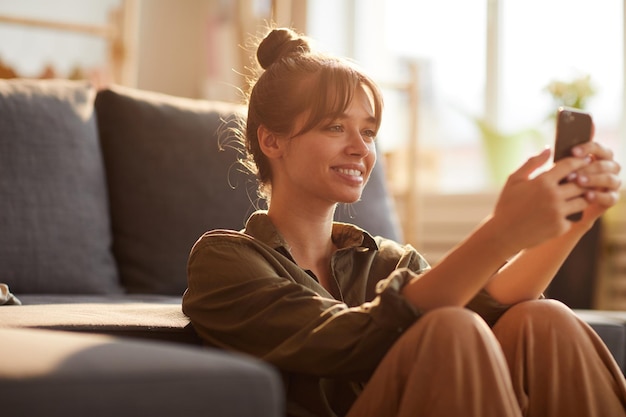 Hermosa chica sonriente con flequillo apoyada en el sofá y navegando por Internet en el teléfono inteligente mientras busca