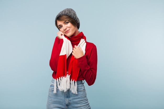 Hermosa chica sonriente con suéter rojo, bufanda y gorro de punto mirando soñadoramente a la cámara sobre fondo azul aislado