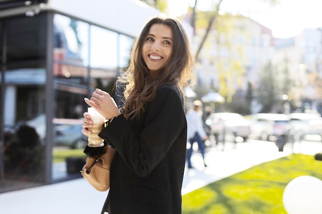 Hermosa chica sosteniendo un vaso de bebida en la calle Atractiva chica bebiendo café al aire libre edificio de cristal en el fondo mirando hacia otro lado