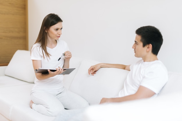 Hermosa chica sostiene un cuaderno con un bolígrafo en la mano y con entusiasmo le hace preguntas a su novio en la moderna sala de estar blanca. Guy escucha atentamente a su novia. Mujer joven sonríe con alegría