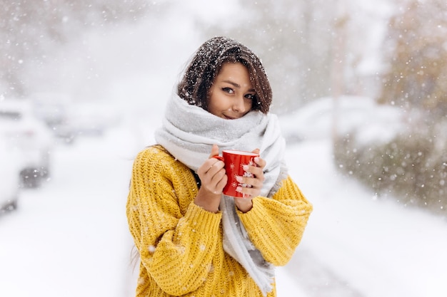 Hermosa chica con un suéter amarillo y una bufanda blanca parada con una taza roja en una calle nevada