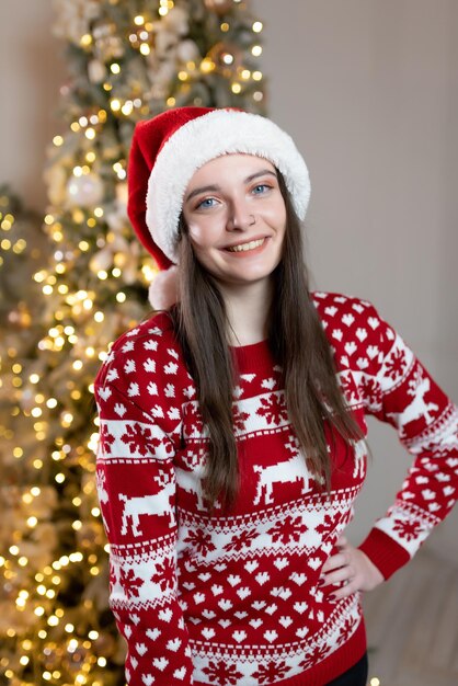 Hermosa chica en un suéter de Navidad y un sombrero rojo de Santa Claus se encuentra cerca de un lujoso árbol de Navidad Celebración sonriente de Navidad de año nuevo