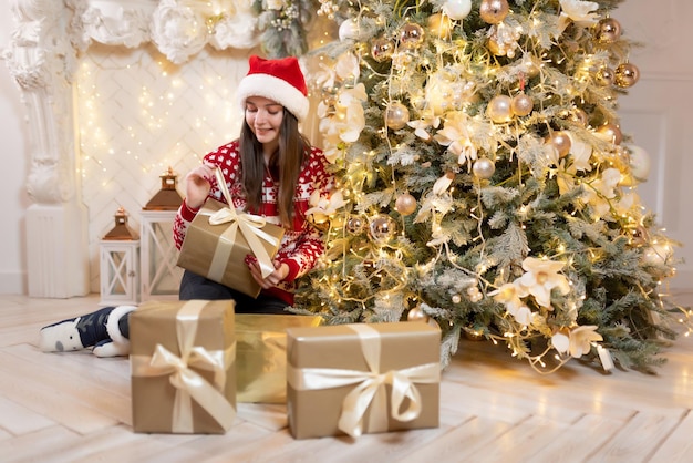 Hermosa chica en un suéter de Navidad y un sombrero rojo de Santa Claus se sienta cerca de un lujoso árbol de Navidad Sonriendo y sosteniendo un regalo Celebración de Navidad de año nuevo