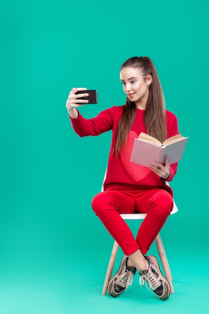 hermosa chica con un suéter rojo lee un libro y usa un teléfono inteligente para selfie