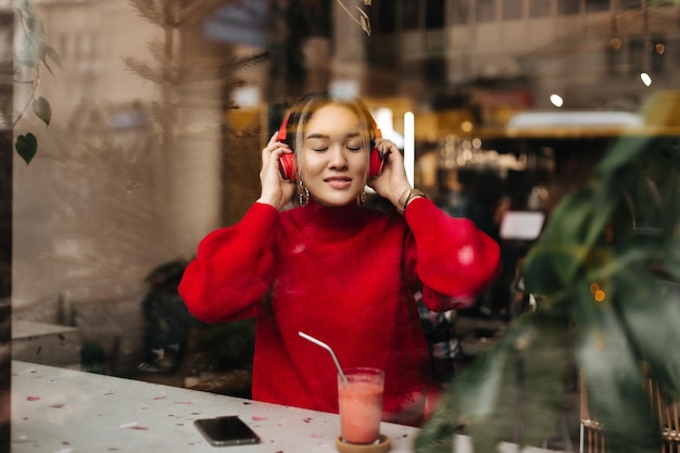 Hermosa chica en suéter rojo se pone grandes auriculares Mujer sentada en el restaurante con batido en la mesa