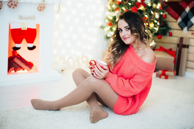 Hermosa chica en suéter rosa, cálido en el fondo de árboles de Navidad y guirnaldas con una taza