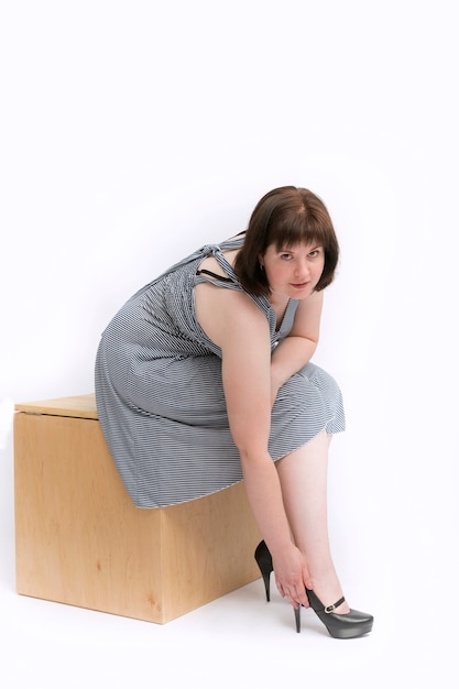 Foto hermosa chica de talla grande con un vestido se sienta en un cajón de madera y endereza sus zapatos de tacón. aislado sobre fondo blanco.