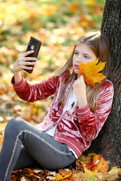 Hermosa chica tomando selfie por teléfono inteligente en el parque de otoño