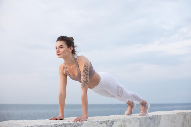 Hermosa chica en top deportivo y polainas blancas practicando yoga con vista al mar en el fondo. Mujer joven mirando soñadoramente a un lado mientras entrena junto al mar