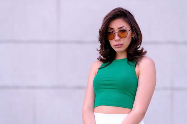 Hermosa chica en un top verde pantalones cortos blancos y gafas de sol posando Mira a la cámara con incredulidad Contra el fondo de una pared gris