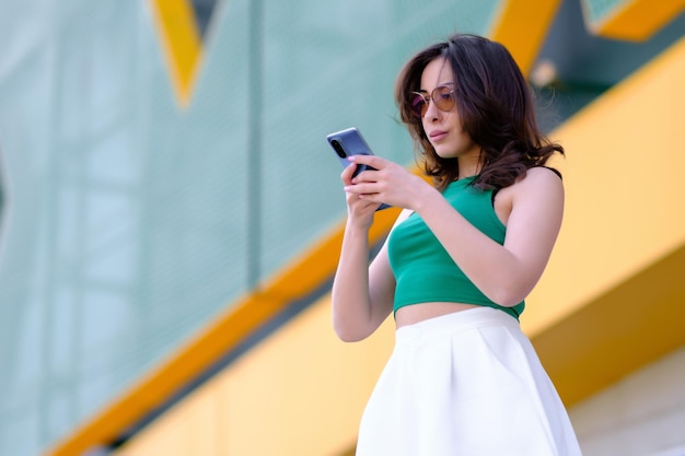 Hermosa chica con un top verde y pantalones cortos blancos sostiene un teléfono móvil en sus manos y lo mira contra el fondo de un edificio amarillo