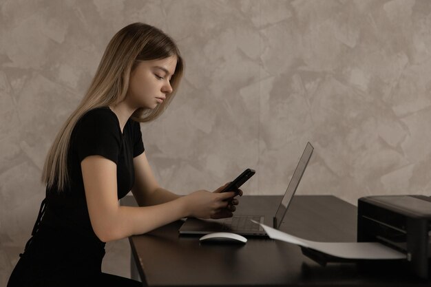 Hermosa chica trabajando remotamente en casa en la computadora y escribiendo un mensaje en el teléfono