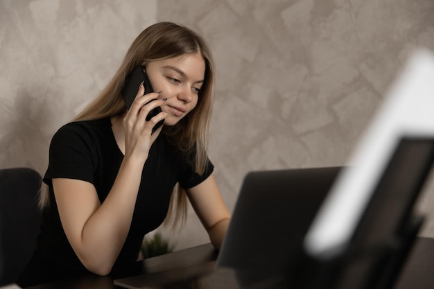 Hermosa chica trabajando remotamente en casa en la computadora y hablando por teléfono