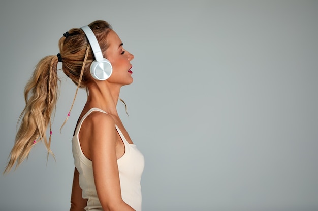 Hermosa chica en traje de baño blanco de moda divirtiéndose sobre fondo gris Increíble mujer rubia sentada en una silla y escuchando música en los auriculares