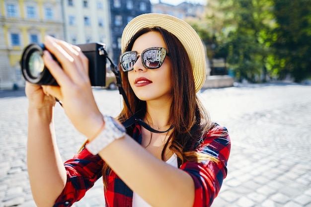 Hermosa chica turista con cabello castaño con sombrero y camisa roja, haciendo fotos con la cámara en el fondo de la antigua ciudad europea y sonriendo, viajando.