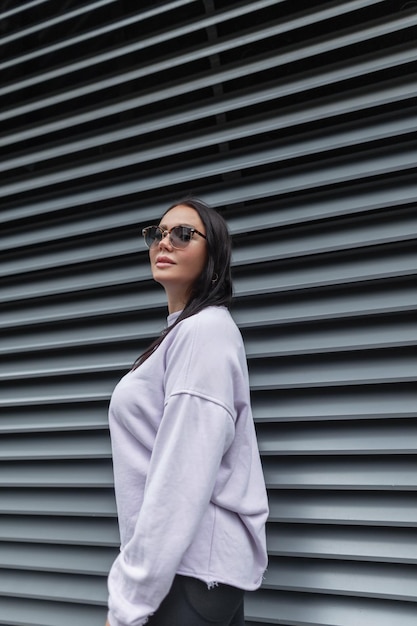 Hermosa chica urbana de moda de talla grande con elegantes gafas de sol en ropa deportiva con un suéter morado cerca de una pared de metal negro con líneas