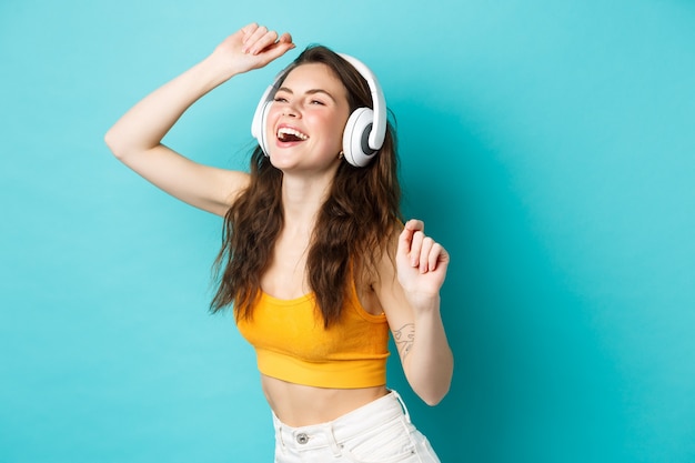 Hermosa chica de verano bailando y riendo en auriculares, escuchando música con una sonrisa despreocupada, de pie sobre fondo azul.