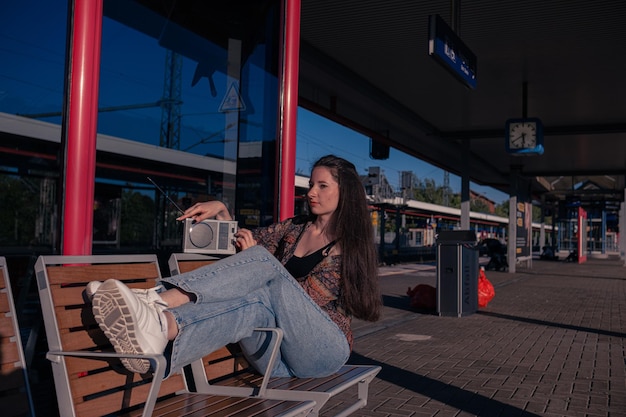 Hermosa chica vestida al estilo de los años 90 con un receptor de radio portátil en sus manos posando en el andén de la estación de tren