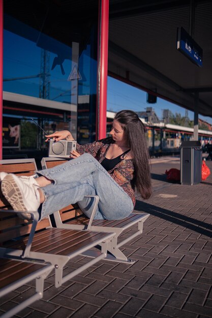 Hermosa chica vestida al estilo de los años 90 con un receptor de radio portátil en sus manos posando en el andén de la estación de tren