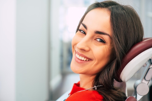 Hermosa chica vestida con un suéter rojo en la sala de estomatología llena de luz del día y colores blancos está sonriendo con su nueva y llamativa sonrisa blanca