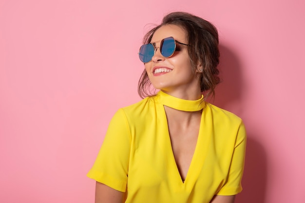 Hermosa chica en vestido amarillo con gafas de sol posando, sonriendo en espacio rosa en estudio