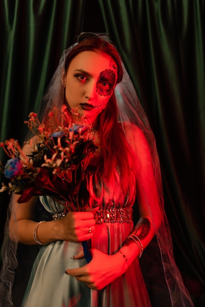 Hermosa chica con un vestido azul con maquillaje artístico en la cara sostiene un ramo de flores Máscara de Halloween Luz roja Concepto de Halloween Mascarada de discoteca