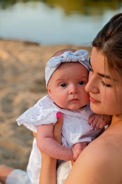 Una hermosa chica con un vestido blanco con un bebé recién nacido está sentada en la playa en la arena