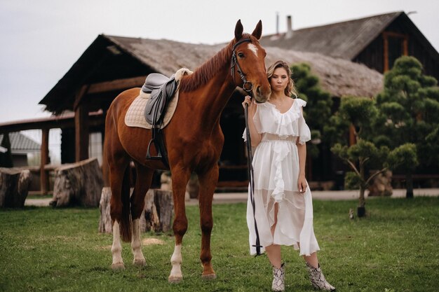 Hermosa chica con un vestido blanco junto a un caballo en un antiguo rancho