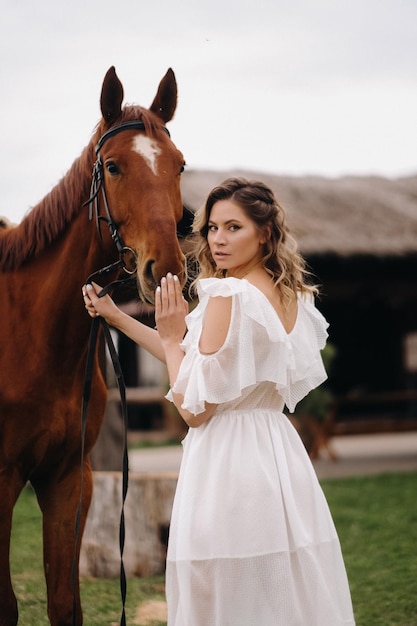 Hermosa chica con un vestido blanco junto a un caballo en un antiguo rancho