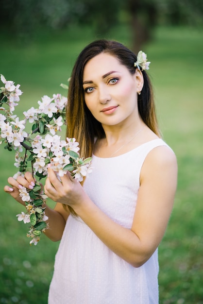 Hermosa chica en vestido blanco con rama de manzano en primavera. Maquillaje profesional, ojos hermosos