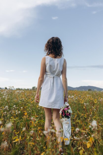 Una hermosa chica con un vestido blanco y un sombrero camina por un camino forestal con flores en las manos El concepto de unas vacaciones de verano