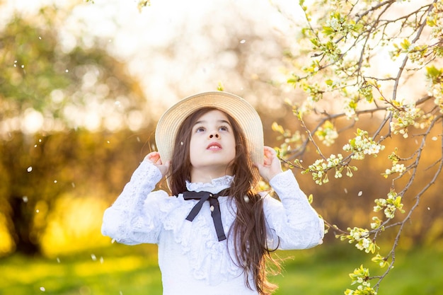 Hermosa chica con un vestido blanco y un sombrero de pie bajo árboles floridos en primavera