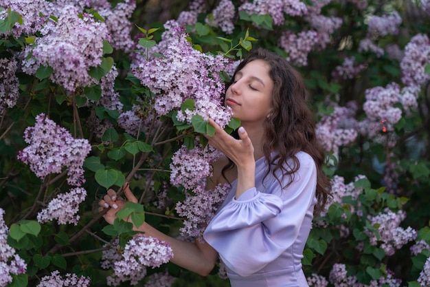 una hermosa chica con un vestido lila está parada cerca de un arbusto respirando el aroma de las lilas