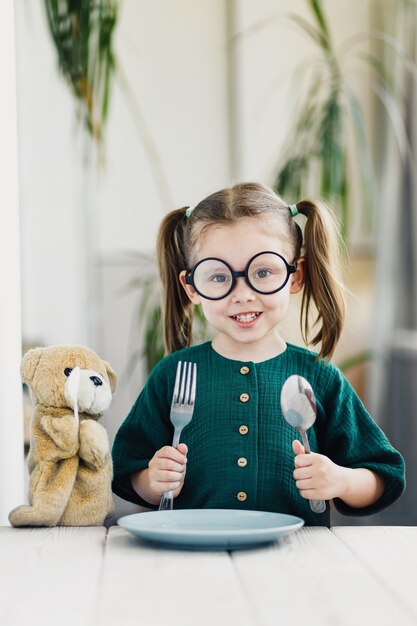 Hermosa chica en vestido de muselina verde esperando el desayuno con oso juguete linda chica en la mesa de comedor