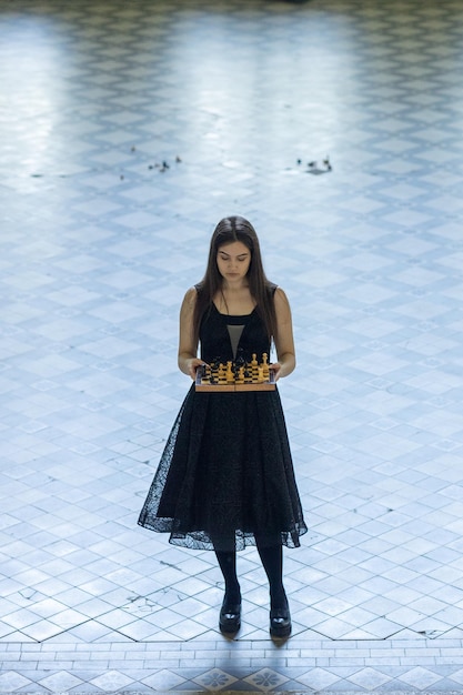 Hermosa chica en vestido negro juega al ajedrez en el piso a cuadros del pasillo