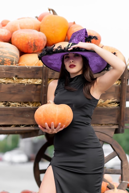 Hermosa chica con un vestido negro y un sombrero de bruja posa cerca de una carretilla llena de varias calabazas Sostiene una calabaza en sus manos Calabaza de Halloween Decoración de calabaza