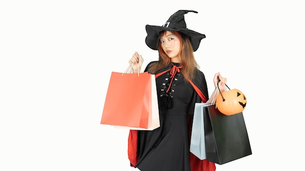 Hermosa chica con vestido negro y sombrero de bruja sostiene bolsas de compras mirando a la cámara y sonriendo en el fondo blanco Concepto de Halloween