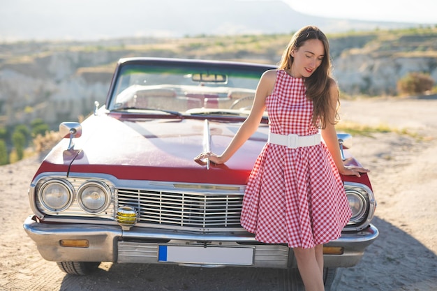 Foto hermosa chica en un vestido retro al amanecer feliz posando en el fondo del paisaje de la montaña en capadocia cerca de un coche retro rojo